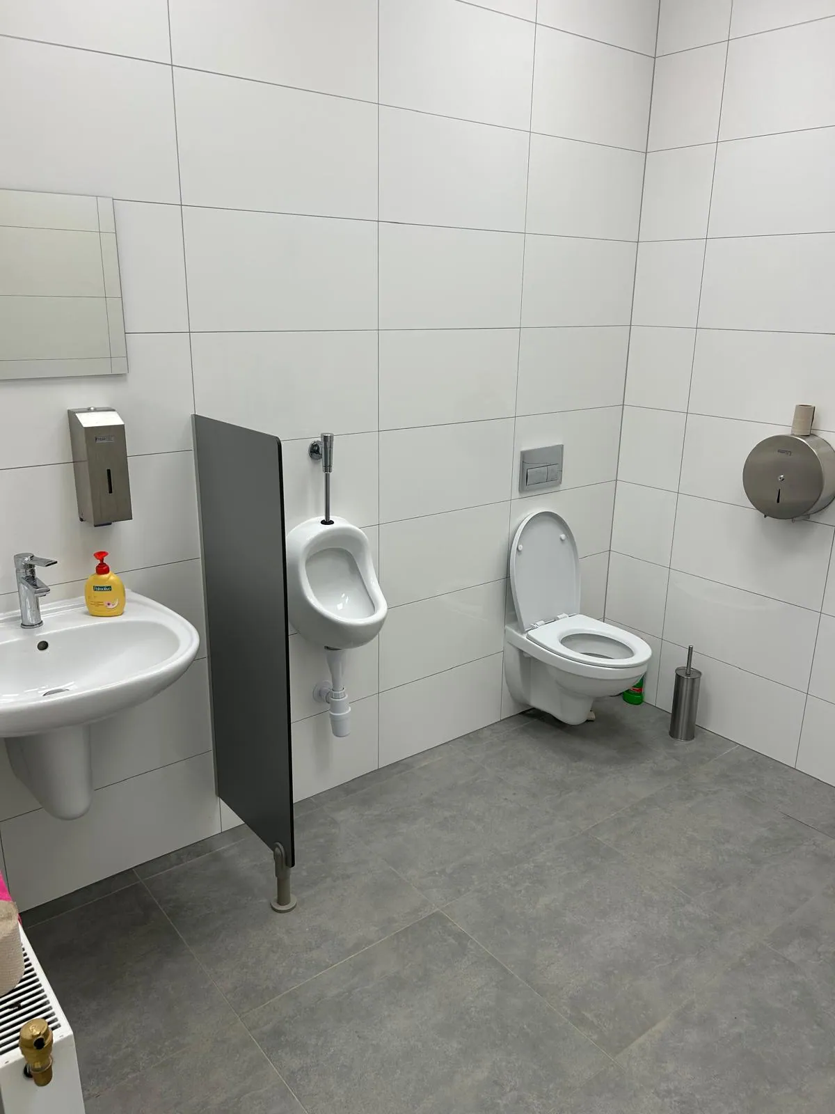 Łazienka z umywalką, pisuarem za przegrodą i podwieszaną miską WC, ściany w białych płytkach